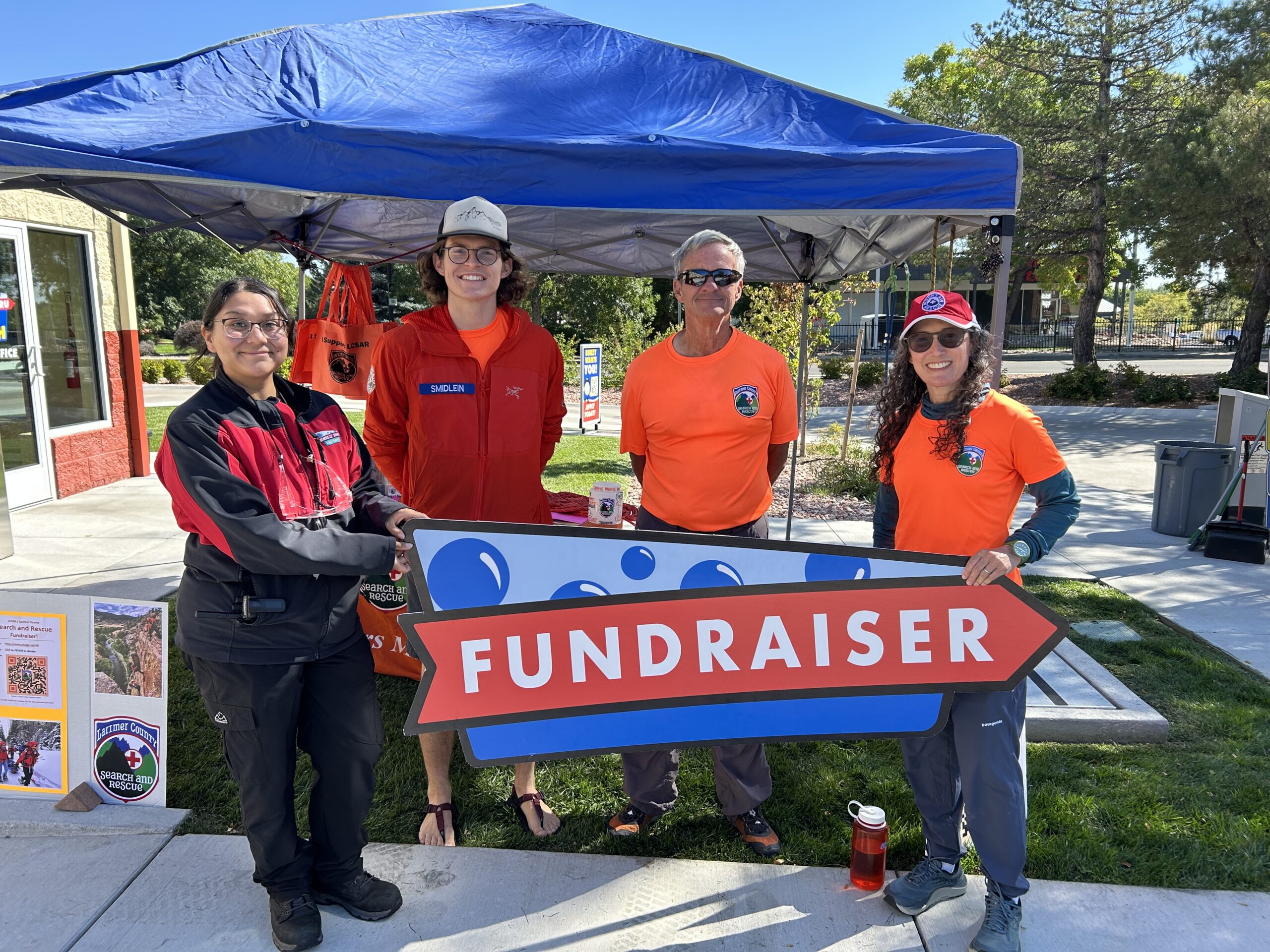 Four people stand outside under a blue canopy, smiling and holding a large "FUNDRAISER" sign for Breeze Thru employment. Two wear orange shirts, one a dark jacket, and one a red jacket. Informational materials are displayed on a table nearby.