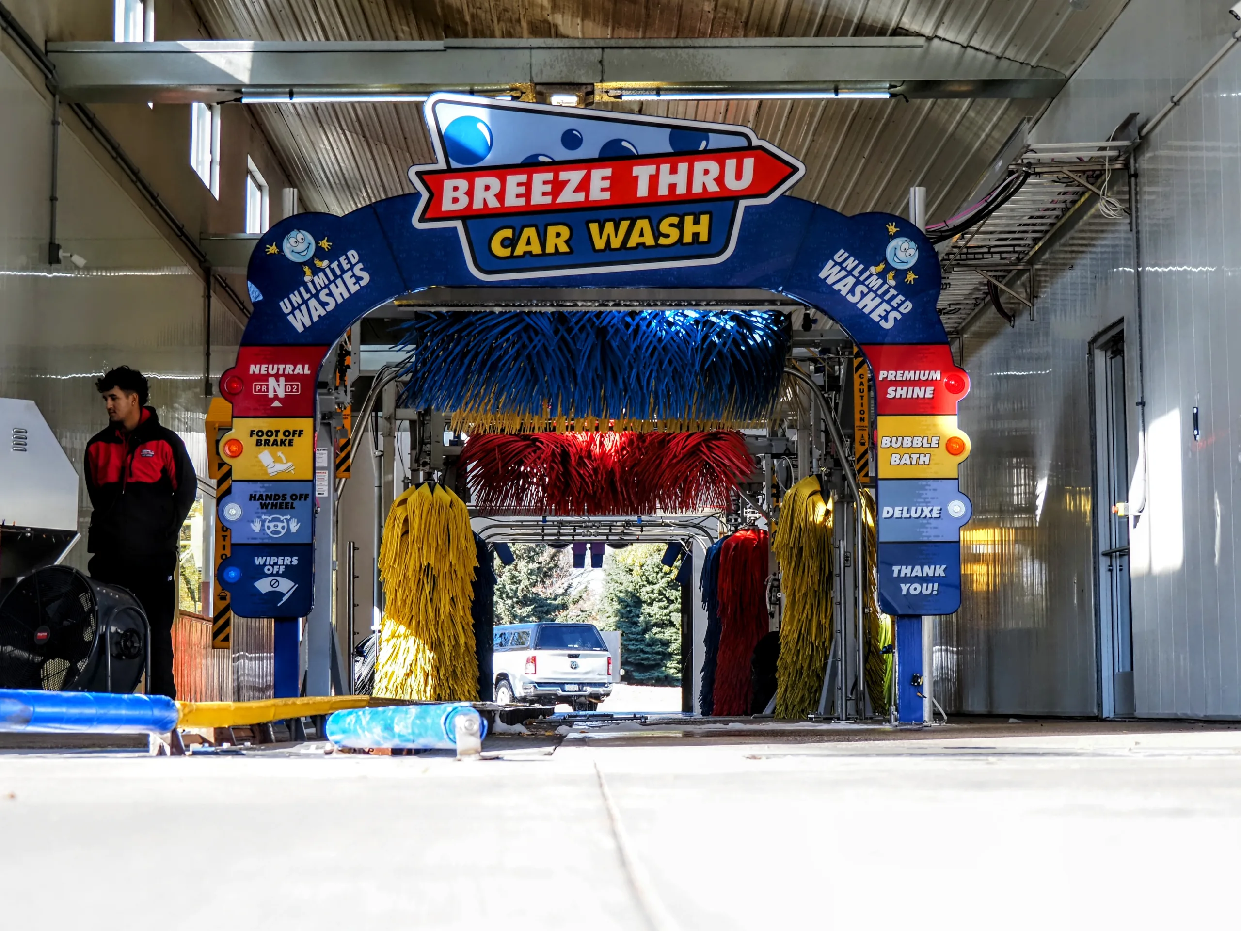 A car enters the Breeze Thru Car Wash, featuring colorful red and blue brushes. Signs indicate different wash options, and a worker stands to the side. The interior is bright and clean.