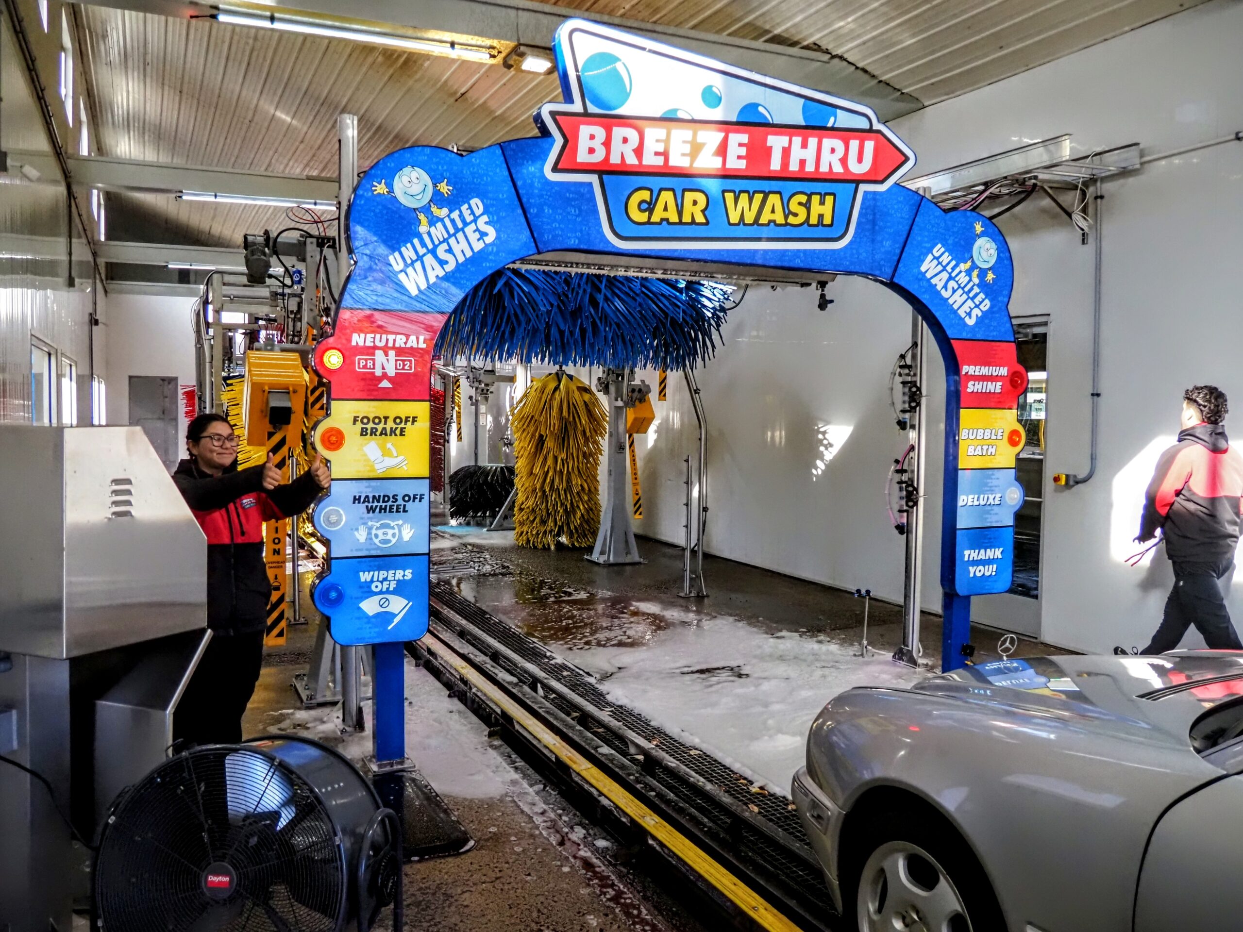 A car enters an automated car wash labeled "Breeze Thru Car Wash." A person in a red jacket stands nearby, operating the system. Another person walks away, and a car is partially visible on the right. The area is brightly lit.