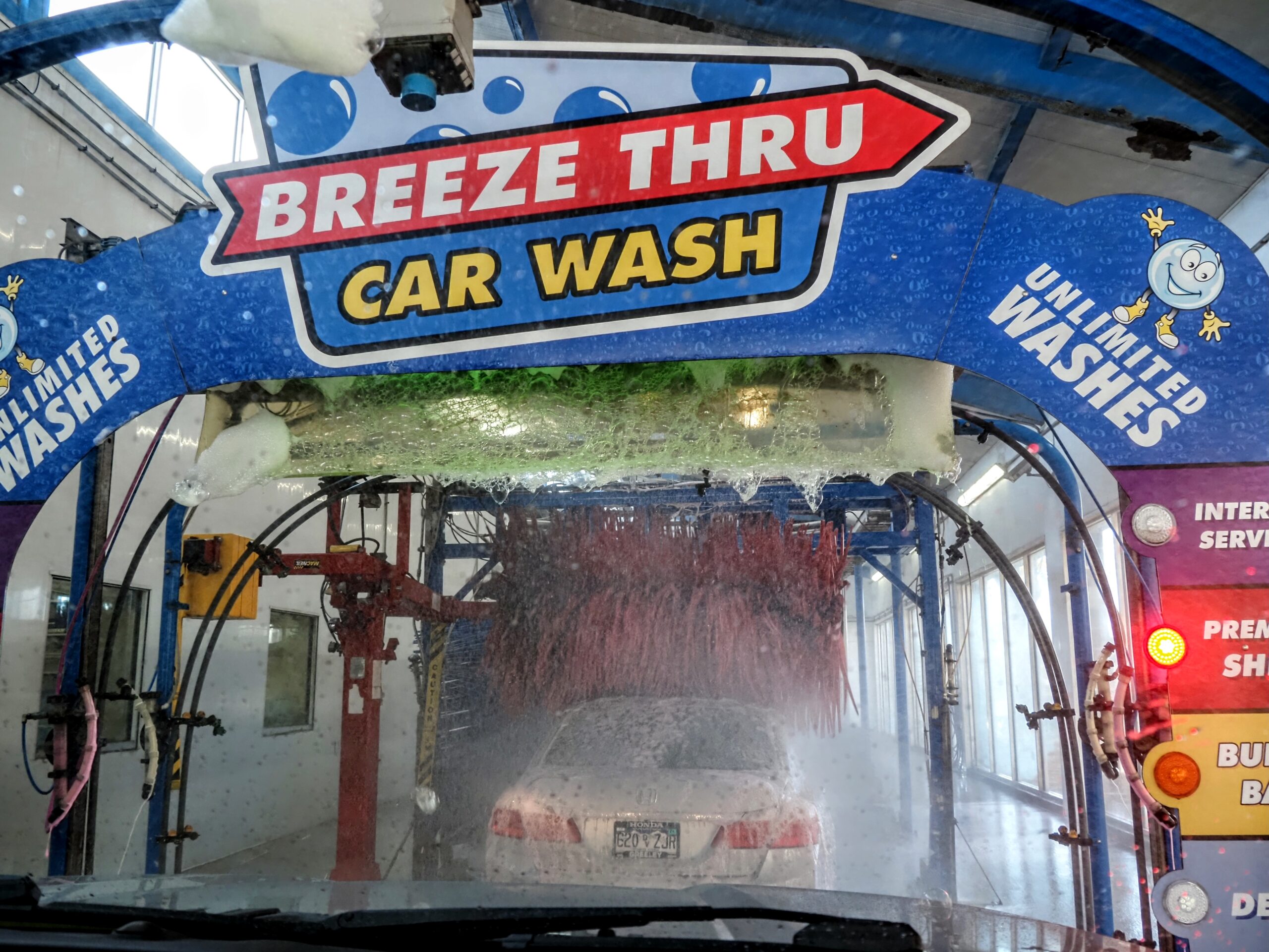 View from inside a car going through a "Breeze Thru Car Wash." The car is surrounded by brushes, soap, and water jets. A sign above reads "Breeze Thru Car Wash" with "Unlimited Washes" banners on each side.