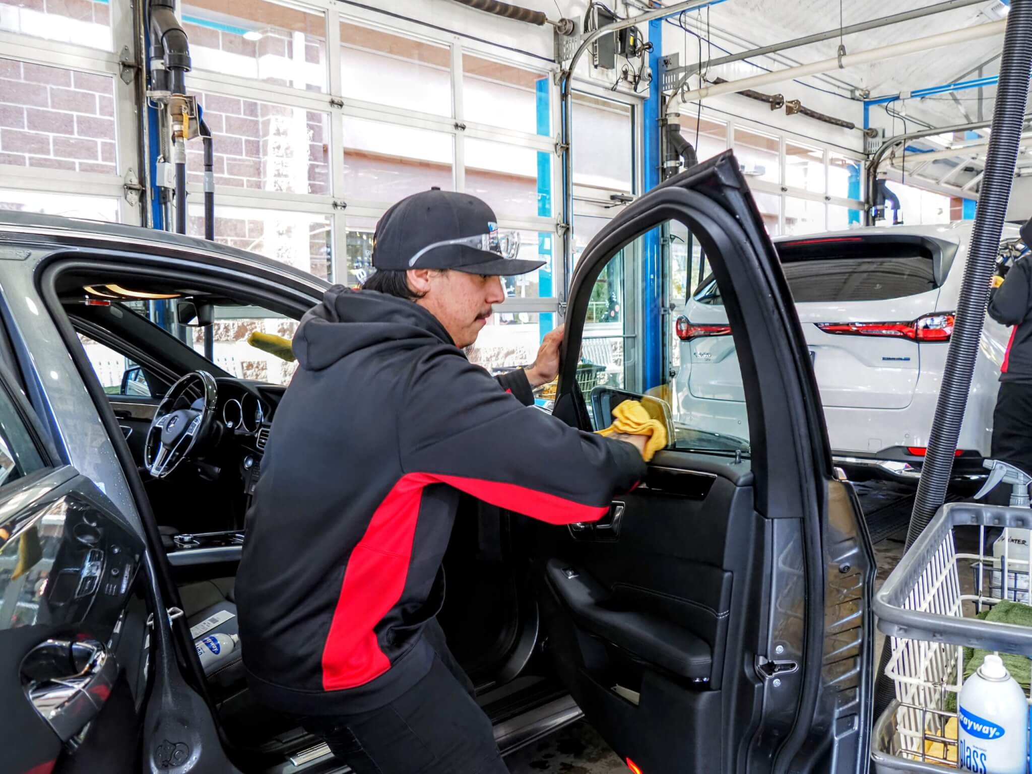 A car wash worker wearing a black cap and red and black uniform is cleaning the inside of a black car door with a yellow cloth. Another vehicle is visible in the background inside the car wash facility. A car wash worker wearing a black cap and red and black uniform is cleaning the inside of a black car door with a yellow cloth. Another vehicle is visible in the background inside the car wash facility.
