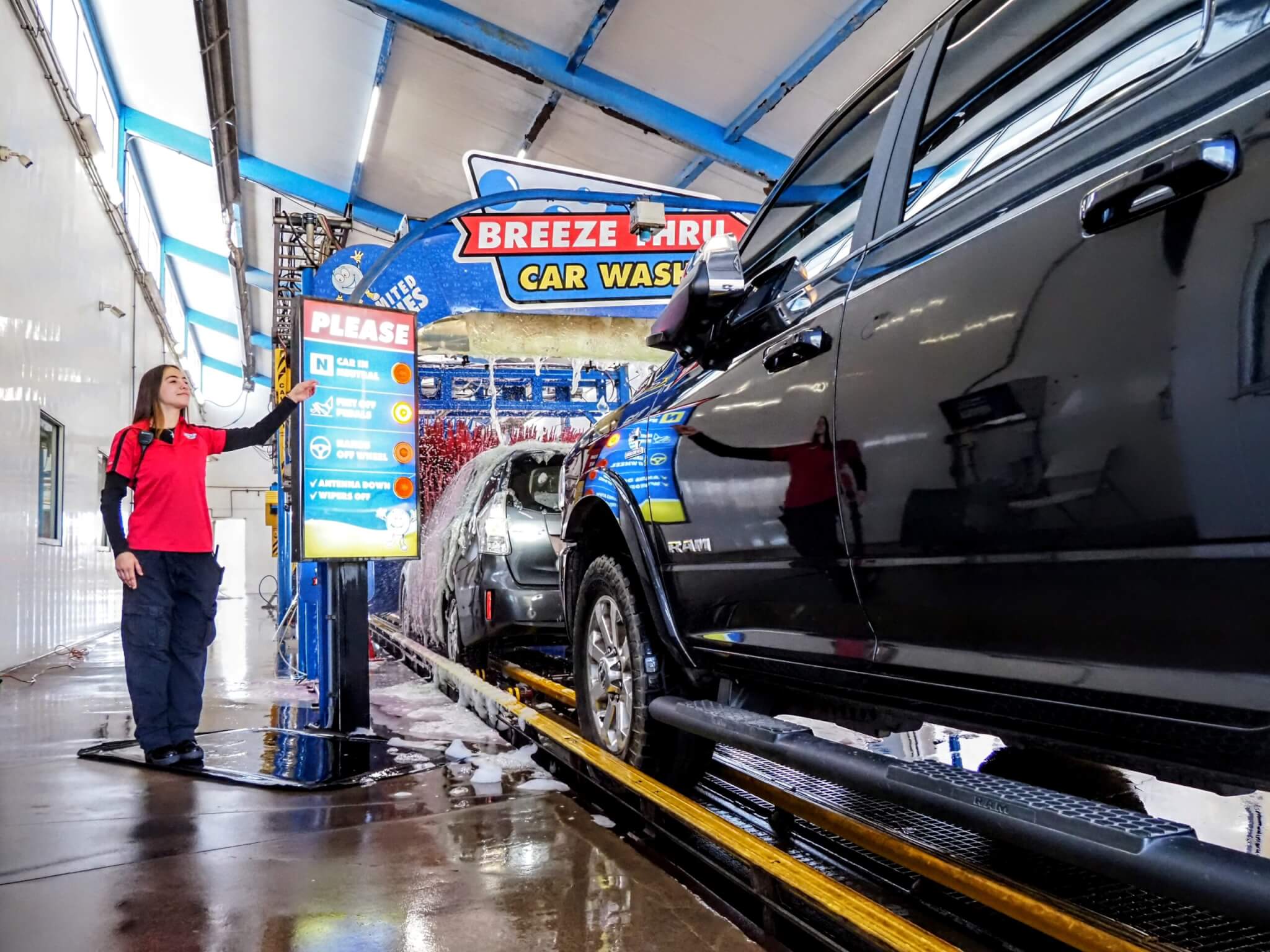 A woman in a red shirt operates a control panel at an automatic car wash. A black pickup truck is on the conveyor belt, moving through the wash, surrounded by soap and water. A woman in a red shirt operates a control panel at an automatic car wash. A black pickup truck is on the conveyor belt, moving through the wash, surrounded by soap and water.