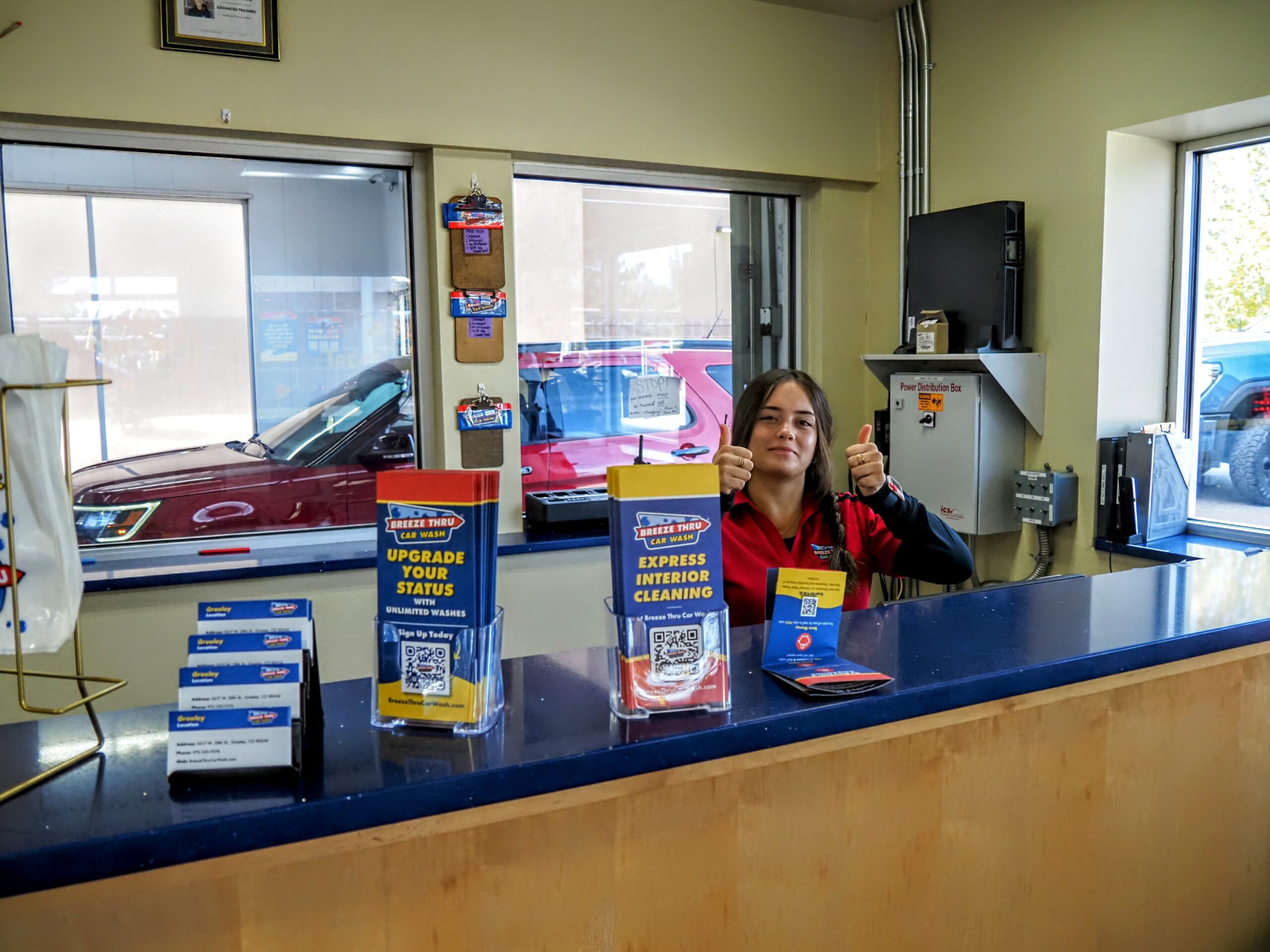 A person stands behind a reception desk inside a car service center, giving a thumbs-up. The desk has brochures and stands with QR codes. A red car is visible through the window on the left.