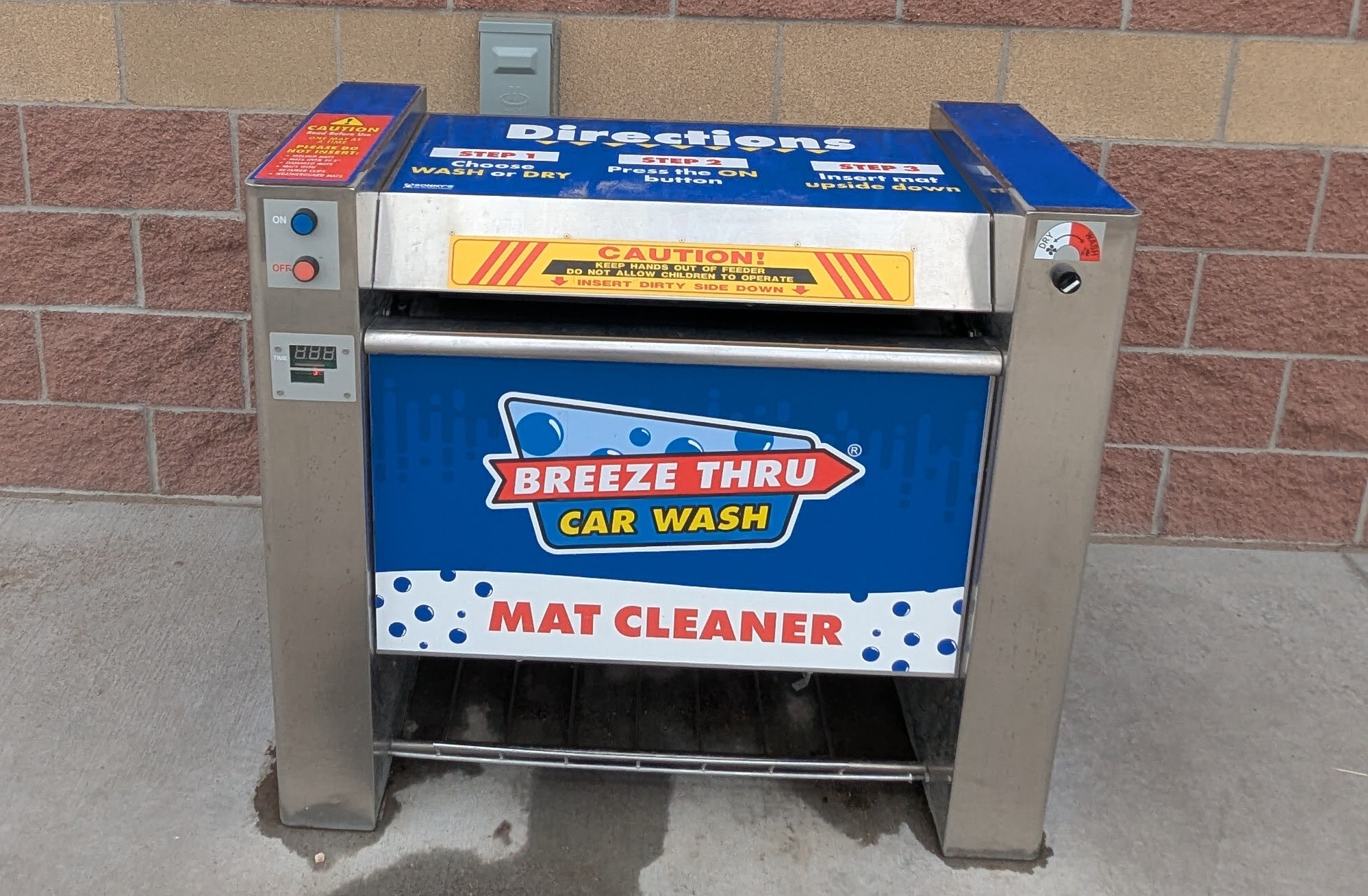 A Breeze Thru Car Wash mat cleaner machine stands outdoors against a brick wall, with buttons, instructions, and caution signs visible on its stainless steel body.