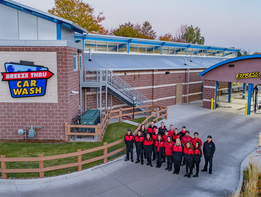 A group of employees in red and black uniforms stand in front of a Breeze Thru Car Wash building and the entrance to the express car wash lane, proudly representing employment opportunities at Breeze Thru.