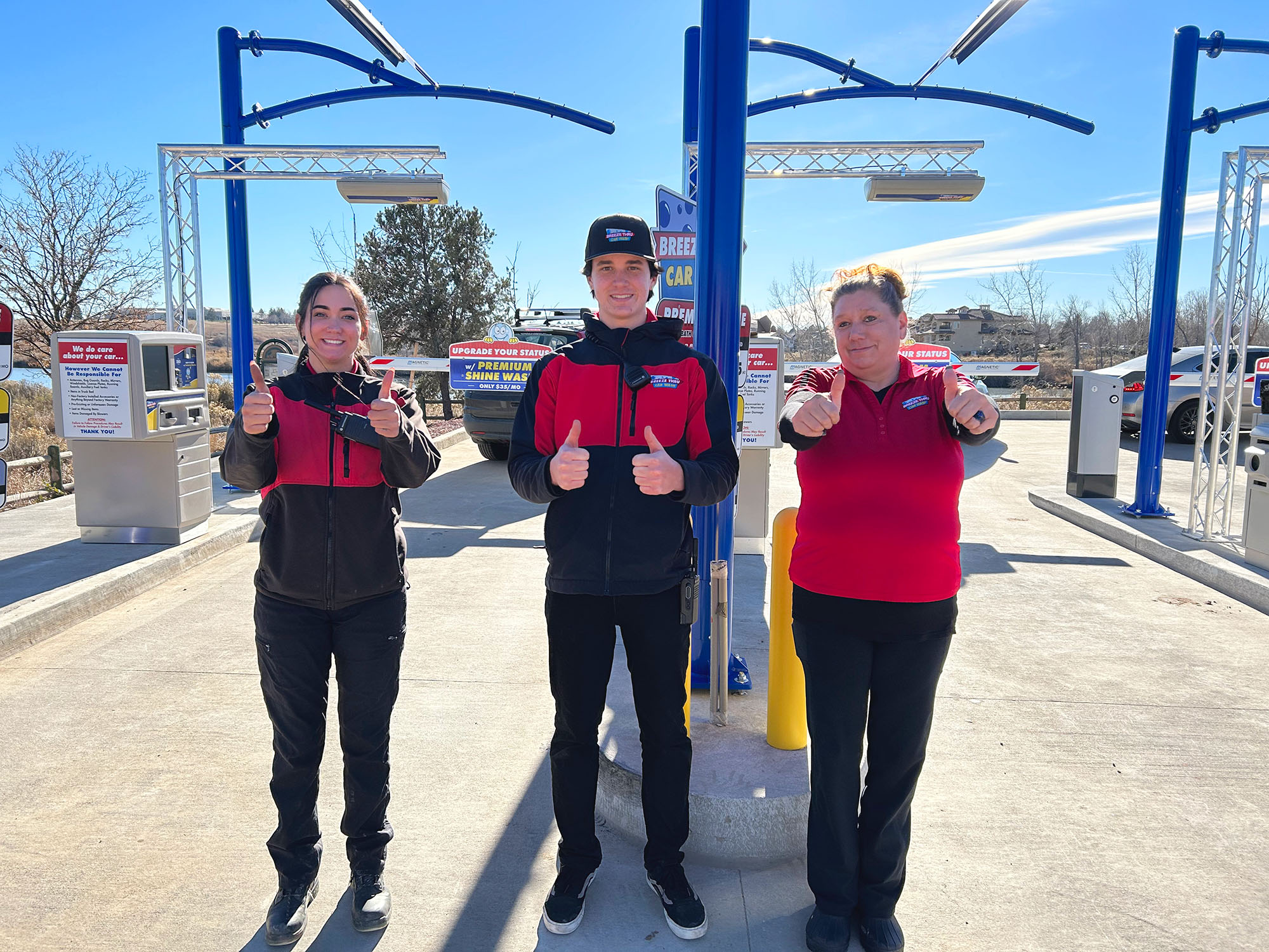 Three car wash employees stand outside in uniform, smiling and giving thumbs up, with car wash equipment and clear blue sky in the background.