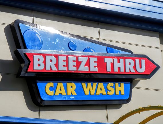 btcw-logo-building A worker in a red and black jacket sprays wax on a dark-colored car inside a car wash. A sign above reads "BUFF IN WAX." The floor is wet, and the car is on a track moving through the wash area.