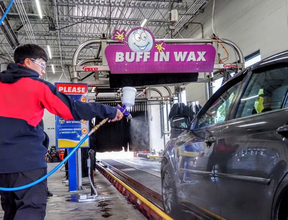 buff-in-wax-spray A worker in a red and black jacket sprays wax on a dark-colored car inside a car wash. A sign above reads "BUFF IN WAX." The floor is wet, and the car is on a track moving through the wash area.