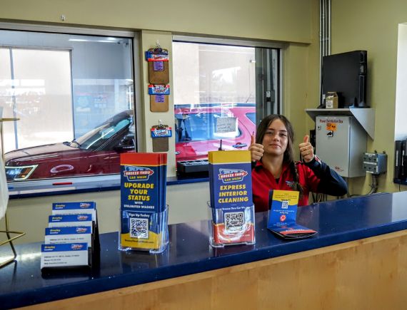 girl-in-office A worker in a red and black jacket sprays wax on a dark-colored car inside a car wash. A sign above reads "BUFF IN WAX." The floor is wet, and the car is on a track moving through the wash area.