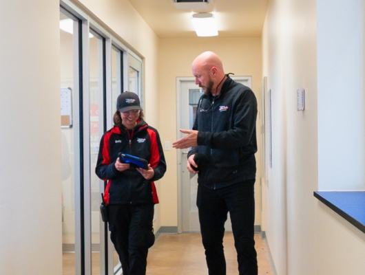 Two people in uniform walk down a hallway, smiling and talking. One holds a blue electronic device, while the other gestures with his hand. The hallway has glass windows and cream-colored walls.